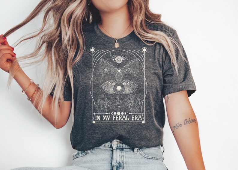 Puede incluir: Una camiseta gris oscuro con un gr&aacute;fico blanco de la cara de un mapache rodeado de estrellas y cuerpos celestes. El texto "IN MY FERAL ERA" est&aacute; impreso debajo del gr&aacute;fico.