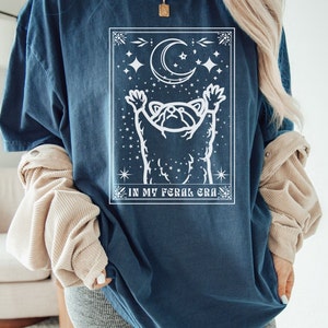 Puede incluir: Una camiseta azul oscuro con un gr&aacute;fico blanco de un mapache que alcanza una luna creciente con estrellas. El texto "IN MY FERAL ERA" est&aacute; impreso debajo del gr&aacute;fico.