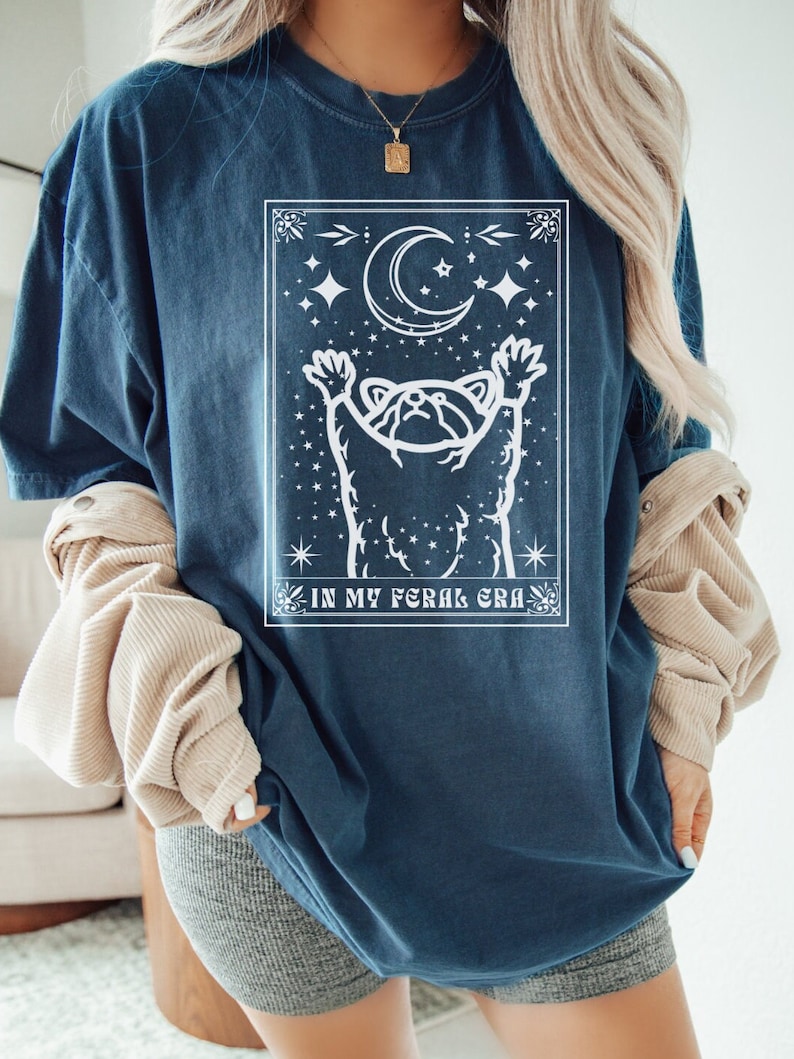 Puede incluir: Una camiseta azul oscuro con un gr&aacute;fico blanco de un mapache que alcanza una luna creciente con estrellas. El texto "IN MY FERAL ERA" est&aacute; impreso debajo del gr&aacute;fico.