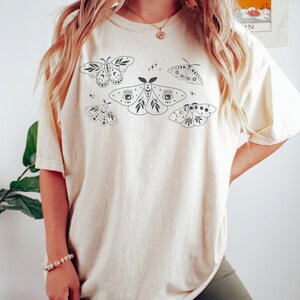 May include: A cream-colored t-shirt with a graphic print of five stylized moths with celestial details. The moths are black and have details like eyes, moons, and stars.