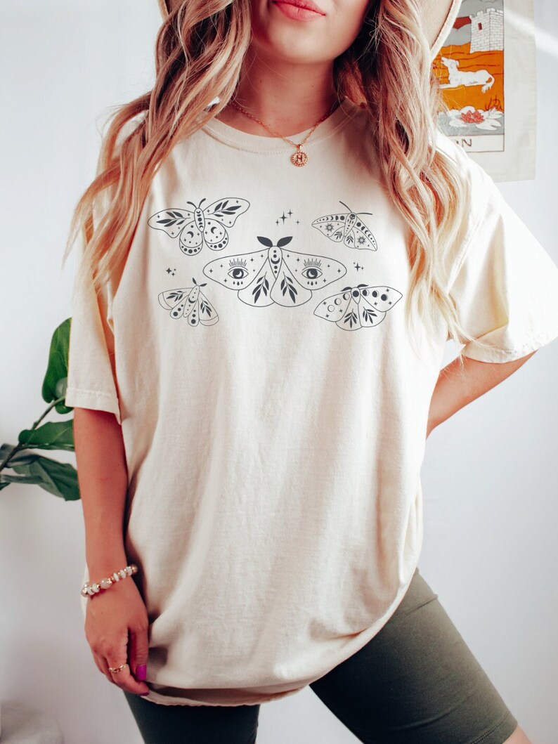 May include: A cream-colored t-shirt with a graphic print of five stylized moths with celestial details. The moths are black and have details like eyes, moons, and stars.