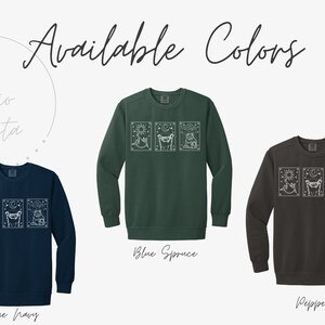 Puede incluir: Se muestran sudaderas en azul marino, verde y gris, cada una con un gr&aacute;fico blanco de tres im&aacute;genes estilo tarot. El texto "Available Colors" est&aacute; en la parte superior, con "Studio Lista" en la esquina superior izquierda. Los colores est&aacute;n etiquetados.