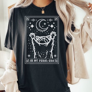 Puede incluir: Camiseta negra con un gr&aacute;fico blanco de un mapache que alcanza una luna creciente con estrellas. El texto "IN MY FERAL ERA" est&aacute; debajo del gr&aacute;fico.