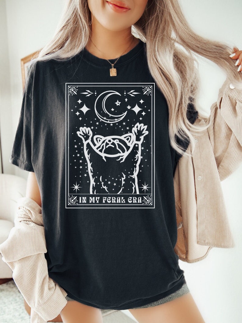 Puede incluir: Camiseta negra con un gr&aacute;fico blanco de un mapache que alcanza una luna creciente con estrellas. El texto "IN MY FERAL ERA" est&aacute; debajo del gr&aacute;fico.