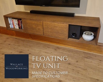 Puede incluir: Un mueble de TV flotante con dos puertas y un estante. El mueble está hecho de madera y tiene un acabado marrón oscuro. El mueble está montado en la pared y tiene un altavoz negro y una maceta blanca con piedras decorativas en el estante. El texto "FLOATING TV UNIT" y "MADE TO CUSTOMER SPECIFICATIONS" se muestra debajo del mueble.