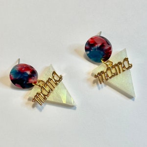 Puede incluir: Un par de pendientes con forma de triángulo blanco y la palabra "mama" en letras doradas. Cada pendiente tiene un charm redondo con estampado azul, rojo y blanco.