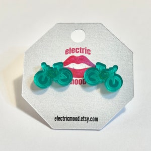 May include: A pair of teal green bicycle earrings on a white card with the text "electric mood" and a red lip graphic. The card also includes the website "electricmood.etsy.com".