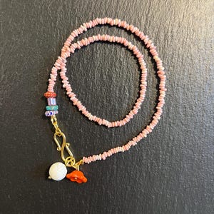 May include: A necklace composed of small, light pink chip beads featuring a gold-toned S-hook clasp. The piece includes a cluster of accent beads in orange, blue, and white, along with a white carved flower charm and an orange flower charm.