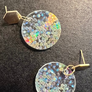 以下が含まれることがあります： 虹色の丸いディスクがついた、金色のイヤリングのペア。ディスクは小さな白いビーズで満たされ、わずかに半透明で、虹色の効果があります。