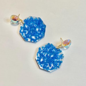 May include: A pair of blue and clear resin earrings with iridescent accents. The earrings are shaped like octagons and feature small blue beads embedded in the resin.