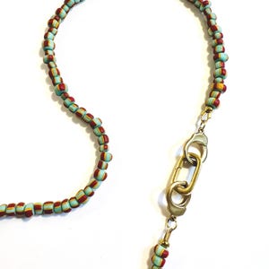 May include: A necklace featuring small, cylindrical beads in alternating stripes of light blue, red, and yellow. The strand is connected by a metallic gold-toned clasp mechanism consisting of two interlocking oval links and a lobster claw closure.
