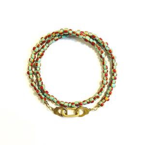 May include: A multi-strand beaded necklace featuring small cylindrical beads in alternating stripes of light blue, red, and yellow. The necklace is secured with a gold-toned metal clasp consisting of two small handcuff-shaped links connected by a central oval ring.
