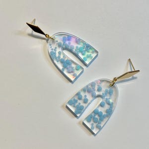 May include: A pair of iridescent blue arch-shaped earrings with gold posts. The earrings are filled with small blue glitter.