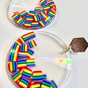 Könnte beinhalten: Klare, runde Ohrringe mit einem goldfarbenen sechseckigen Stecker. Die Ohrringe haben regenbogenfarbene quadratische Perlen, die in klarem Harz eingeschlossen sind. Die Perlen sind im Inneren des klaren, kreisförmigen Ohrringdesigns angeordnet.