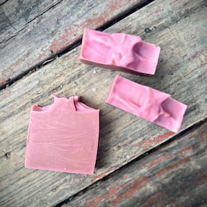 May include: Three rectangular bars of solid pink soap with a smooth, matte finish and textured, swirled tops. The soap bars are arranged in a cluster, displaying a consistent dusty rose color throughout each piece with subtle variations in the surface.