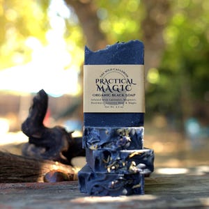 May include: A stack of four rectangular black soap bars with visible dried herbs embedded in the texture. A tan paper label wraps around the top bar, printed with the text The Wild Cauldron Practical Magic Organic Black Soap, Net Wt. 4.5 oz.