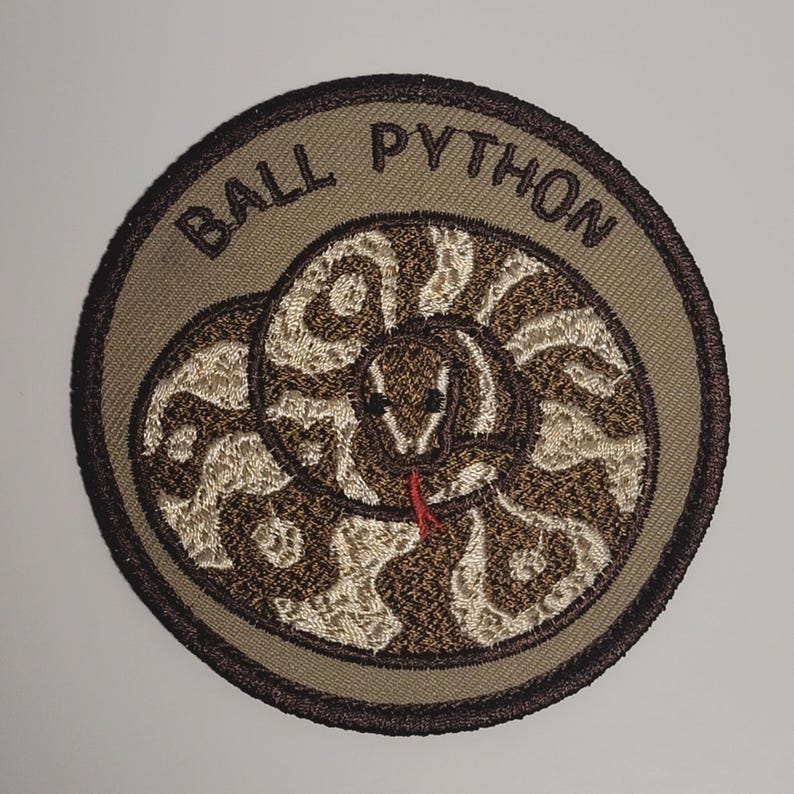 May include: A tan and brown embroidered patch featuring a white ball python coiled around itself. The patch has the text "BALL PYTHON" in brown embroidery around the snake.