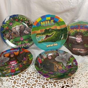 Puede incluir: Colección de cinco platos coloridos con ilustraciones de animales de la jungla. Un plato tiene un elefante, otro un cocodrilo con el nombre "NILE", y un tercero con un orangután llamado "OZZIE". Los platos son de "Rainbow Cafe".