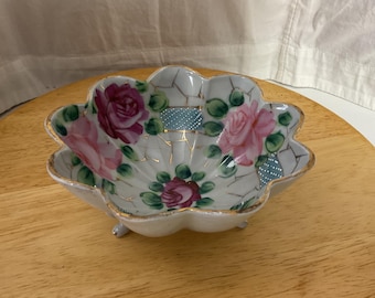May include: A white porcelain bowl with a scalloped edge and three feet. The bowl is decorated with pink and red roses, green leaves, and gold accents. The bowl has a cracked ice design.