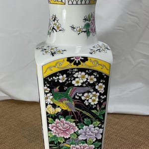May include: A white ceramic vase with a rectangular body and a flared top. The vase is decorated with a colorful design featuring a bird, flowers, and geometric patterns in yellow, pink, green, and black.