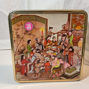 May include: A vintage square tin with a gold-coloured frame. The lid features a colourful illustration of a traditional Asian banquet scene with numerous figures, food, and decor. The image includes text in a pink circle.