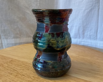 Könnte beinhalten: Eine kleine, keramische Vase mit einer einzigartigen, mehrfarbigen Glasur. Die Vase hat eine bauchige Form mit einem schmalen Hals und einem breiten Boden. Die Glasur zeigt Farbtöne von Grün, Blau, Rot und Braun, wodurch ein marmorierter und strukturierter Effekt entsteht.