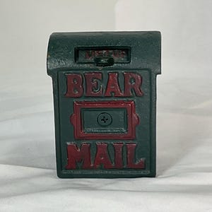 Pode incluir: Uma caixa de correio de parede em ferro fundido verde-escuro com o texto vermelho em relevo BEAR MAIL na frente. A caixa retangular tem uma tampa superior articulada com uma ranhura e um painel central retangular fixado por um parafuso.