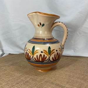 Op de afbeelding: Een crèmekleurige keramische kan met een decoratief bloemmotief. De kan heeft een handvat en een tuit, met blauwe en oranje banden rond de body. Het bloemmotief bevat bruine, groene en gele elementen.