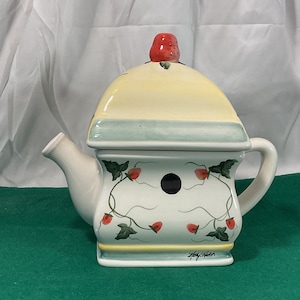 May include: A white ceramic teapot with a yellow and green birdhouse design. The teapot has a red strawberry on the lid and a black hole on the front. The teapot is signed "Holly Hobbie".