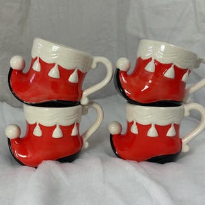May include: Four ceramic mugs shaped like red elf boots with white trim and handles. The mugs are stacked in two pairs, showcasing their festive design. Each boot has a white ball on the toe.
