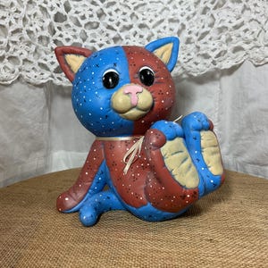 May include: A ceramic cat figurine with a patchwork design. The cat is painted in blue and red, with white polka dots. It has large black eyes, a pink nose, and beige ears. The cat is sitting with its paws outstretched.