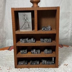 May include: A wooden display shelf with multiple compartments. The shelf contains metal letterpress type, including the words "OLD", "THEIR", and "DRIVE". A small framed image of a golfer is also present. The shelf has a wooden knob on top.