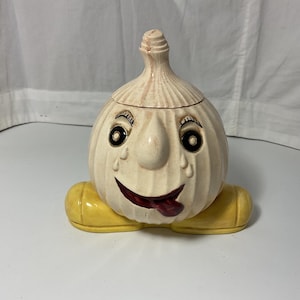 Pode incluir: Um recipiente de cerâmica caprichoso em forma de alho com tampa. O recipiente tem um rosto sorridente com olhos pretos, uma língua vermelha e pés amarelos. O alho é esbranquiçado com ranhuras verticais.