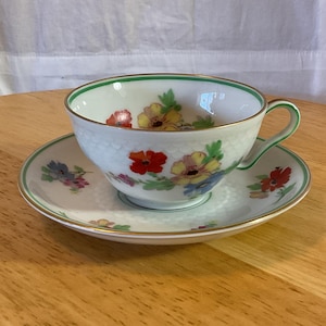 Op de afbeelding: Een wit theekopje en schotelset met een bloemenpatroon. Het kopje heeft een groene rand en de schotel heeft een gouden rand. De bloemen zijn rood, geel, blauw en groen.