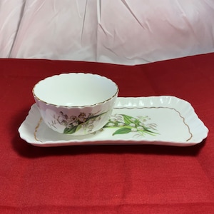 May include: A white porcelain bowl with a gold rim and a floral design sits on a matching white tray with a gold rim. The tray and bowl feature a lily of the valley design.