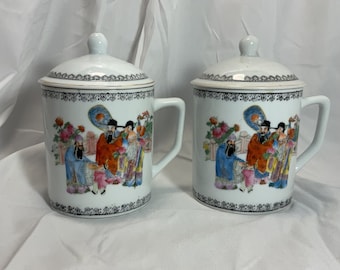 Puede incluir: Dos tazas de cerámica blanca con tapas, que presentan un diseño chino colorido de figuras con ropa tradicional. Las tazas tienen un borde negro y detalles dorados.