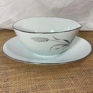 May include: White gravy boat and saucer set with a silver trim. The gravy boat features a wheat stalk design in gray. The set is on a brown burlap surface.