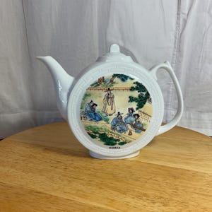May include: White ceramic teapot with a round design featuring a scene of people in traditional Korean clothing. The teapot has a handle and spout and says "KOREA" on the bottom.