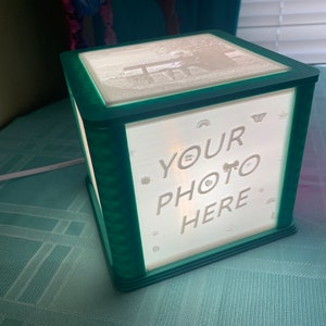 May include: A teal blue cube-shaped light box with a white frosted panel. The panel has a white image of a person sitting on a bench and the text "YOUR PHOTO HERE" with small decorative elements.