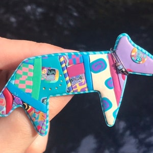 Könnte beinhalten: Ein farbenfroher Hund-Anstecker aus Polymer Clay. Der Anstecker hat ein Patchwork-Design mit verschiedenen Mustern und Farben, darunter Türkis, Rosa, Lila und Creme. Eine silberne Nadel sichert die Brosche. Der Hund hat einen skurrilen, künstlerischen Stil.