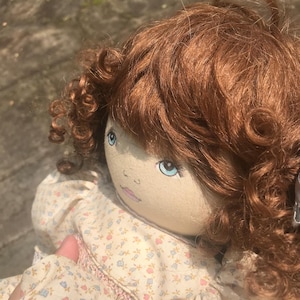 May include: A handmade doll with a fabric face, blue eyes, and auburn curly hair. The doll wears a cream-colored dress with a floral pattern and pink trim. The doll is a vintage style.