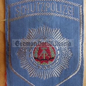 Könnte beinhalten: Ein blauer Stoff-Patch mit silberner Stickerei. Der Patch zeigt den Text "SCHUTZPOLIZEI" und einen silbernen Stern mit roter und goldener Mitte.