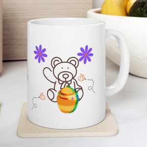 May include: White ceramic mug with a brown line drawing of a teddy bear holding a honey pot. The bear is wearing a bow tie and there are purple flowers and bees on the mug.