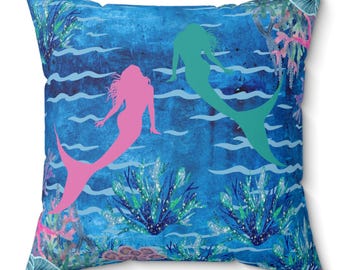 May include: Square throw pillow featuring a vibrant underwater scene. Two mermaid silhouettes, one pink and one teal, swim amidst blue water, coral, and aquatic plants. The pillow has a blue background with a textured appearance.