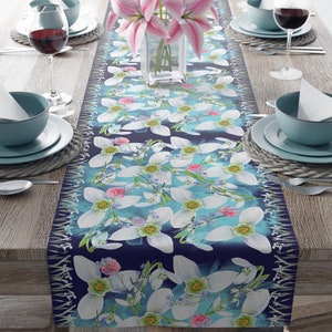 May include: A blue and white floral table runner with a pattern of white flowers and green leaves. The runner is on a wooden table with two place settings on each side.