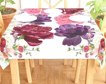 May include: A white rectangular tablecloth featuring a floral design with large, watercolor-style peonies in shades of burgundy, pink, and purple. Green leaves and smaller flowers accent the design. The tablecloth is set on a wooden table.
