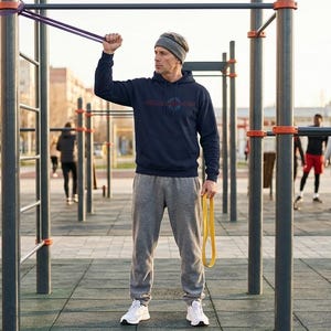 Könnte beinhalten: Eine Person, die einen marineblauen Hoodie mit Logo, graue Jogginghose und eine graue Mütze trägt und im Freien mit Widerstandsbändern trainiert. Die Trainingsgeräte sind schwarz mit orangefarbenen Akzenten.