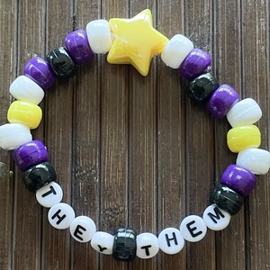 May include: A yellow star charm bracelet with black, white, purple, and yellow beads. The bracelet spells out "They Them" in black letters on white beads.