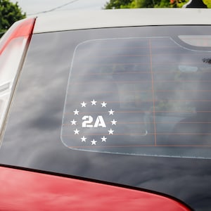 以下が含まれることがあります： 白いビニール製の車のステッカーで、星の円の中に「2A」の文字が描かれています。ステッカーは赤い車のリアウィンドウに貼られています。テキストと星は、明確で太字のフォントです。