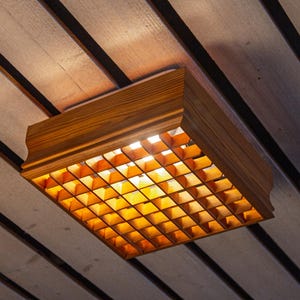 May include: A square wooden ceiling light fitting featuring a grid-patterned lattice shade. The light emits a warm, golden glow through the open square compartments of the wooden frame, which is mounted directly against a panelled ceiling surface.